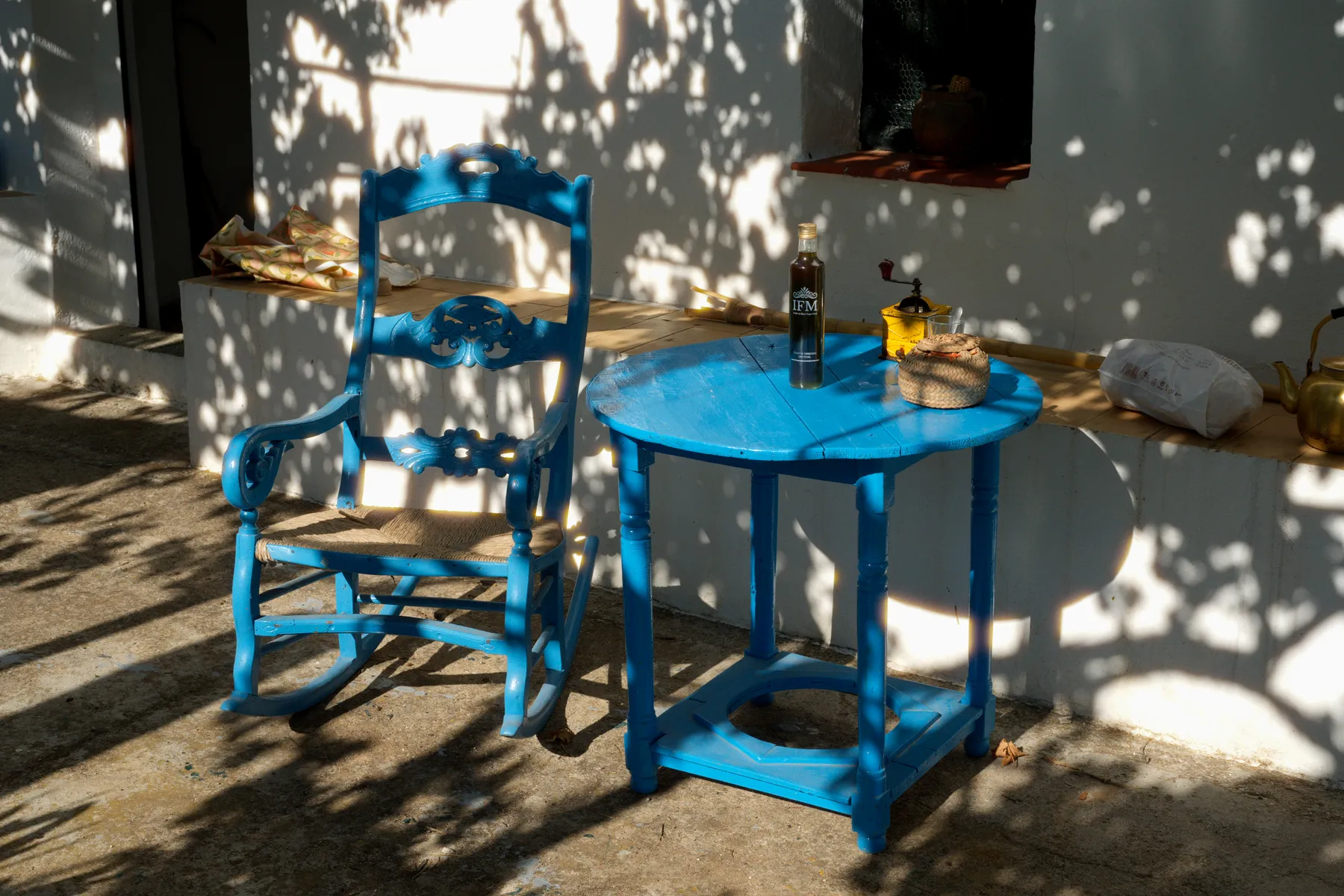 Una silla y una mesa azules junto a una pared encalada, a la sombra de un árbol.